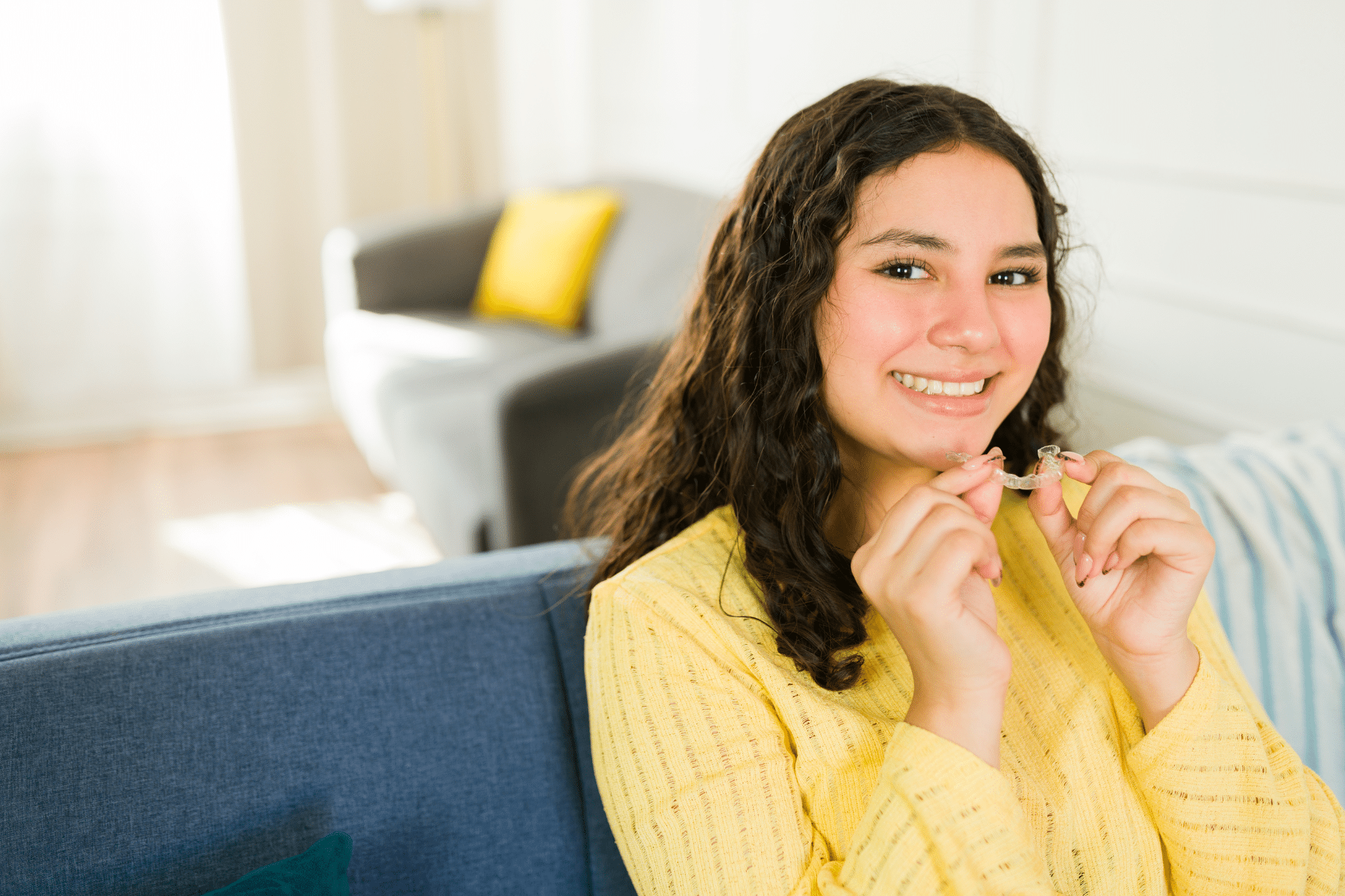 Smiling young woman holding a clear aligner while sitting on a couch